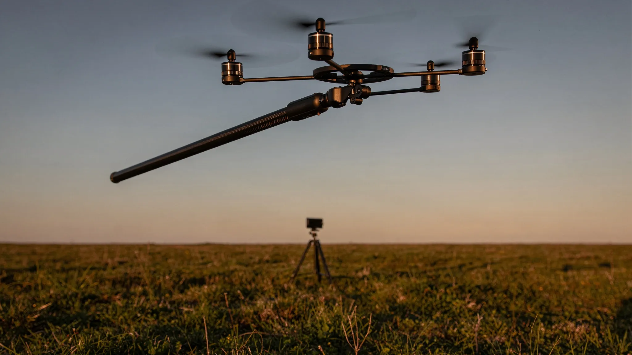A thin carbon-fibre pole robot with four small propellers at its top, caught in mid-air above a grassy field, a small tripod in the distance, in warm evening light.