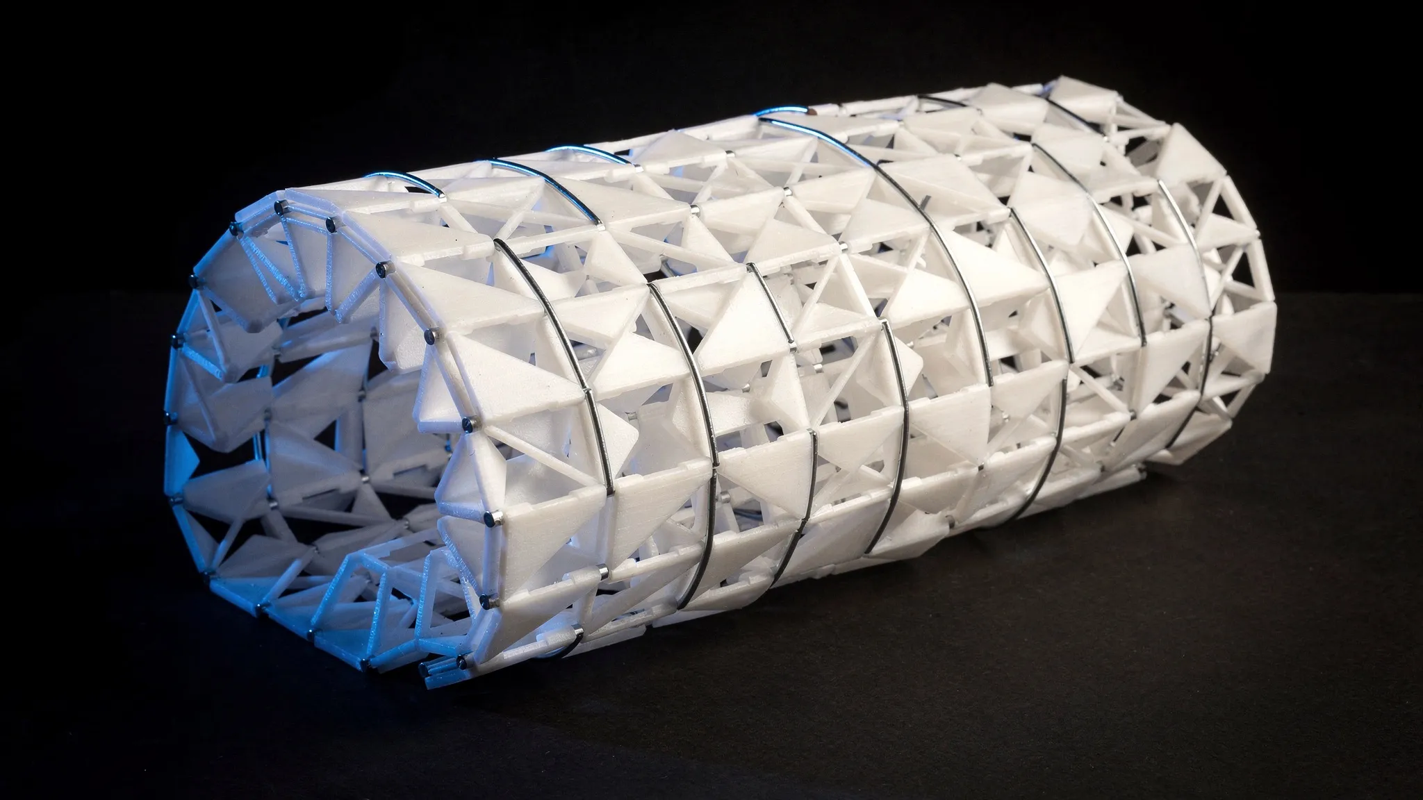 A sheet of small white triangular panels folded into a cylinder, thin steel strips arched across its hinges, lit in blue.