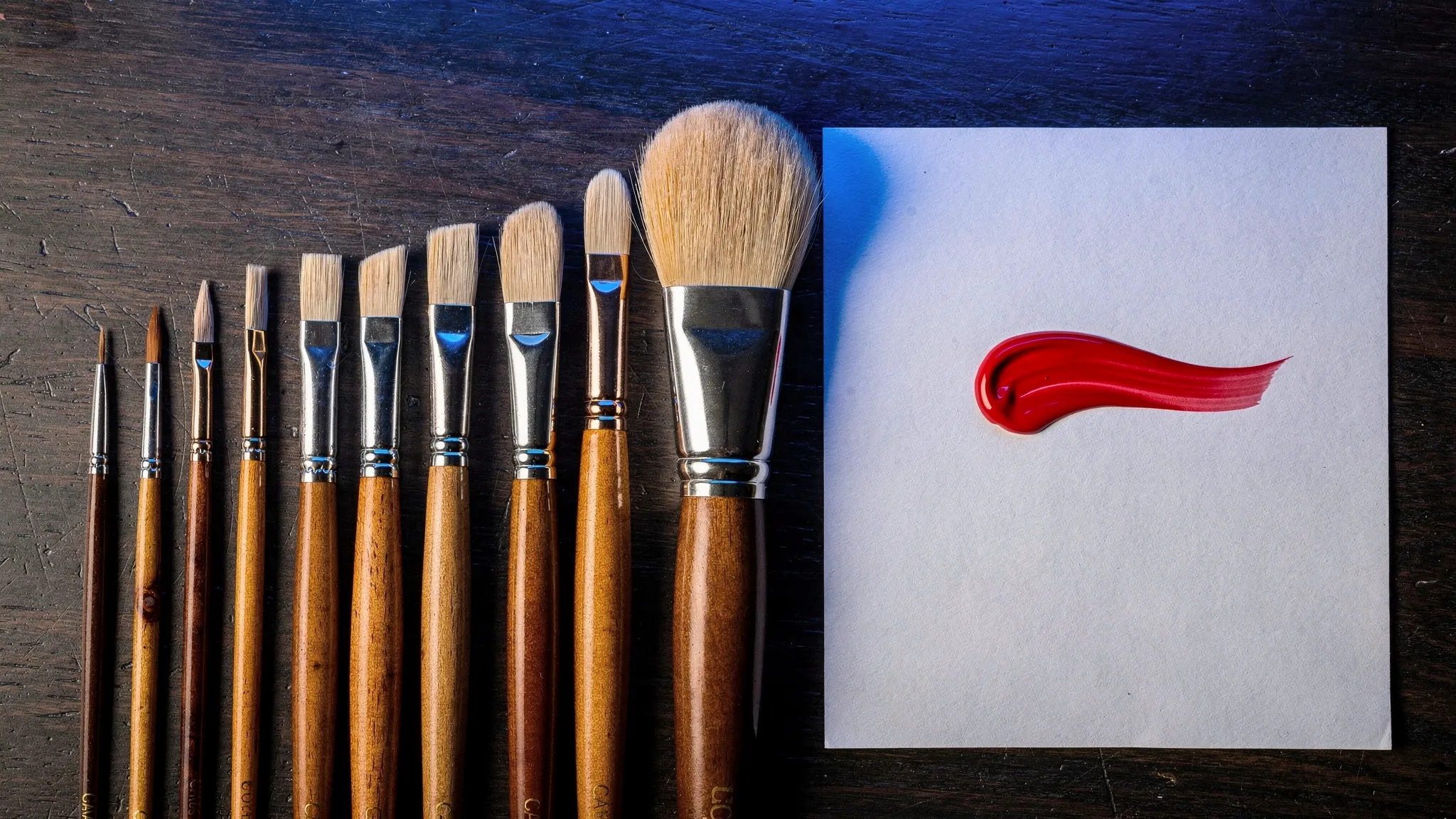 A row of paintbrushes of steadily increasing size lying side by side on a dark table, a red paint stroke on white paper beside them, lit in deep cobalt blue.